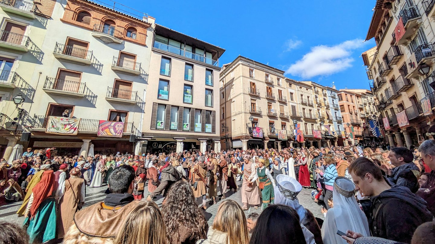 People in historical costumes in a sunny town square with banners.
